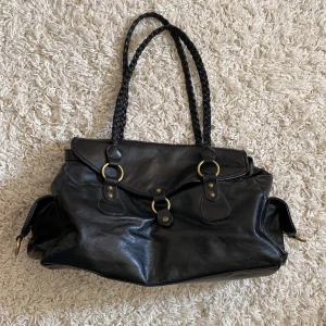 Svart skinnväska med flätade handtag - Snygg svart skinnväska med flätade handtag och guldfärgade detaljer. Väskan har en praktisk design med flera fack och stängs med knappar. Perfekt för att bära med sig allt man behöver under dagen.