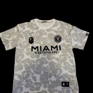Vit och grå kamouflage fotbollströja från BAPE - Säljer en cool oanvänd vit och grå kamouflage fotbollströja från BAPE med 'MIAMI' och 'A BATHING APE' på framsidan. Tröjan har korta ärmar och ett stort 'BAPE 93' tryck på baksidan. Perfekt för sommaren skriv om ni vill se fler bilder( pris kan diskuteras) 