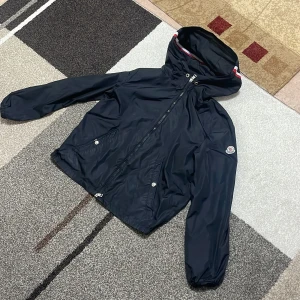 Svart vindjacka från Moncler - Snygg svart vindjacka från Moncler med dragkedja och huva. Jackan har långa ärmar och två fickor med knappar framtill. Perfekt för blåsiga dagar och har en stilren design med Moncler-logga på ärmen.