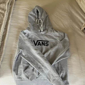 Grå hoodie från Vans - Säljer en stilren ljuslila/grå hoodie från Vans med svart logga på bröstet. Tröjan har en klassisk känguruficka och justerbar huva med dragsko. Perfekt för en avslappnad stil.