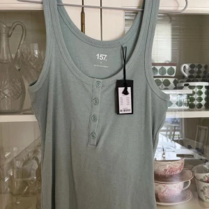 Grön ribbad linnetopp från 157 - Säljer en stilren grön ribbad linnetopp från 157. Toppen har knappar framtill och är tillverkad i en mjuk blandning av bomull och polyester. Perfekt för en avslappnad look! En lite prick på kanten (se bild), vart där från köp! 