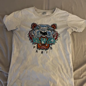 Äkta kenzo t shirt  - Säljer en vit t-shirt från Kenzo med ett coolt tigertryck i färgglada nyanser på framsidan. T-shirten har korta ärmar och en rund halsringning. Perfekt för en avslappnad stil med en touch av lyx.
