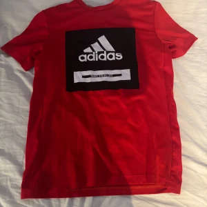 Röd t-shirt från Adidas - Säljer en röd t-shirt från Adidas med en stor svart och vit logga på framsidan. T-shirten är kortärmad och tillverkad i ett lätt och bekvämt material, perfekt för aktiva dagar. Adidas Aeroready-teknologi för extra komfort. Passar ungdomar i storlek 13-14 år.