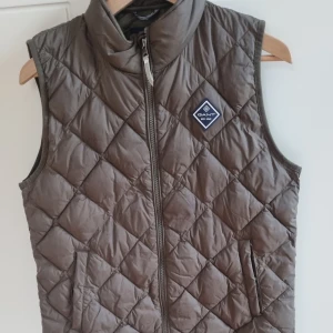 Mörk grönquiltad väst från GANT - Säljer en snygg brun quiltad väst från GANT. Västen har en dragkedja framtill och ett stilrent märke på bröstet. Perfekt för lager-på-lager under kyligare dagar. Passar ungdomar i åldern 13-14 år passar mig som har s perfekt och är 178cm.