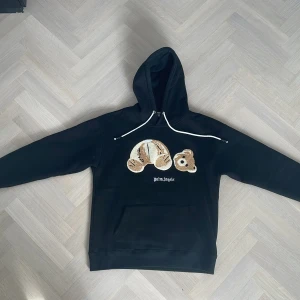 Svart hoodie från Palm Angels med nallemotiv - Säljer en svart hoodie från Palm Angels med ett unikt nallemotiv på framsidan. Tröjan har en klassisk huva med vita dragsnören och en stor ficka framtill. Storlek L. Perfekt för en avslappnad stil. ‼️INTE 0RIGINELL‼️