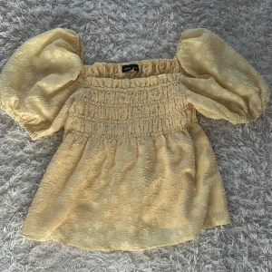 Gul blus från LMTD - Charmig gul blus från LMTD med puffiga korta ärmar och smockdetaljer. Perfekt för en somrig look med sitt blommiga mönster och luftiga design.
