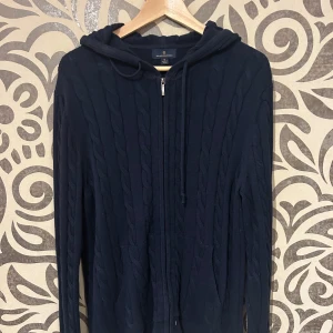 Mörkblå stickad hoodie med dragkedja från Brooks Brothers - Säljer en snygg mörkblå stickad hoodie med dragkedja framtill. Tröjan har en klassisk kabelstickad design och en praktisk huva med snörning. Skriv för fler bilder, org pris 1400kr