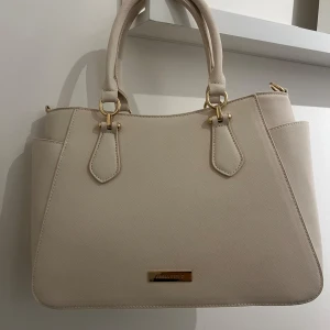 Beige handväska från Anna Field🌸 - Elegant beige handväska från Anna Field med stilrena detaljer i guld. Väskan har två handtag och en praktisk dragkedja upptill. Insidan är rymlig med flera fack för enkel organisering. Perfekt för att bära med sig det nödvändigaste med stil.❤️❤️ säljs då jag aldrig fått en användning för den❤️