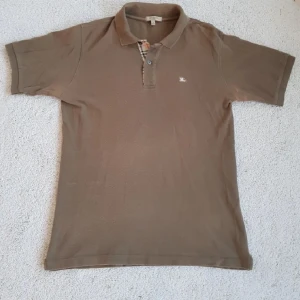 Beige pikétröja från Burberry - Säljer en klassisk beige pikétröja från Burberry i storlek S. Tröjan är tillverkad i 100% bomull och har en stilren krage med knappar. Detaljerad med Burberrys ikoniska rutmönster vid knappslån och en liten broderad logotyp på bröstet.