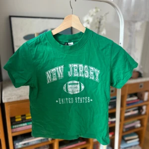 Grön t-shirt med New Jersey-tryck - Säljer en grön t-shirt från Gildan med tryck av 'New Jersey' och en fotboll. Perfekt för sportiga tillfällen eller som en casual vardagströja. Kortärmad och i mjukt bomullsmaterial.