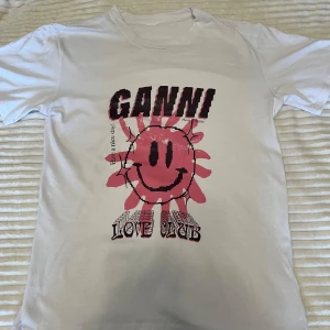 Vit t-shirt med tryck från Ganni - Säljer en vit t-shirt från Ganni med ett coolt tryck av en rosa smiley och texten 'Love Club'. Perfekt för en avslappnad stil. T-shirten har korta ärmar och en normal passform. Skicka ett meddelande vid intresse!