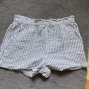 Randiga shorts från Kappahl - Säljer ett par randiga shorts från Kappahl i blått och vitt. De har en bekväm resårmidja med snörning och är perfekta för varma dagar. Shortsen har en avslappnad passform och är tillverkade i ett lätt material.