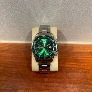 Klocka med grön urtavla och silverarmband - Snygg klocka med grön urtavla och silverarmband. Klockan har tydliga siffror och markeringar samt en datumvisare. Perfekt för den som vill ha en stilren och iögonfallande accessoar.