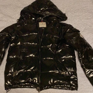 Svart pufferjacka från Moncler - Snygg svart pufferjacka från Moncler med glansig finish. Jackan har en huva och dragkedja framtill samt två fickor med dragkedjor. Perfekt för kyligare väder. Även bra och ha när det blir höst/ vår.                                    Storlek S