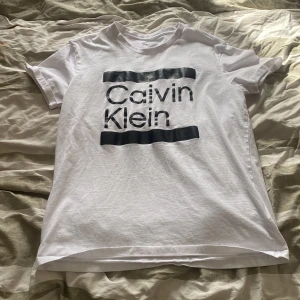 Vit t-shirt från Calvin Klein - Säljer en stilren vit t-shirt från Calvin Klein med deras ikoniska logga i svart på framsidan. T-shirten är kortärmad och gjord i mjuk bomull. Andvänd 2-3 gånger