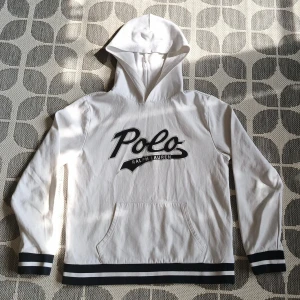 Vit hoodie från Ralph Lauren - Säljer en stilren vit hoodie från Ralph Lauren med svart logotyp på framsidan. Tröjan har långa ärmar och en praktisk magficka. Perfekt för en avslappnad stil.