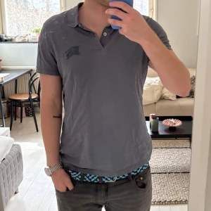 Grå pikétröja med broderad logga - Säljer en stilren blå pikétröja från Burberry med en diskret broderad logga på bröstet. Tröjan har korta ärmar och en klassisk krage med knappar. Perfekt för en avslappnad look.