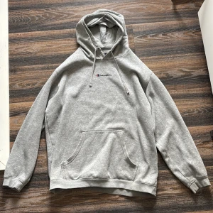 Grå hoodie från Champion - Säljer en klassisk grå hoodie från Champion med broderad logga på bröstet och ärmen. Tröjan har en stor ficka framtill och justerbar huva med dragsko. Perfekt för en avslappnad stil.