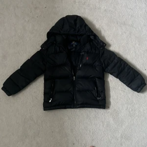 Svart dunjacka från Ralph Lauren - Säljer en snygg svart dunjacka från Ralph Lauren med avtagbar huva. Jackan har dragkedja och två fickor med dragkedjor framtill. Den klassiska röda loggan syns på bröstet. Perfekt för kyliga dagar!