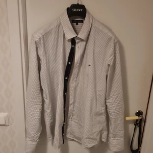 Randig skjorta från Tommy Hilfiger - Säljer en stilren randig skjorta från Tommy Hilfiger i storlek XL. Skjortan är tillverkad i 97% bomull och 3% lycra, vilket ger en bekväm passform. Den har långa ärmar och klassiska knappar framtill. Perfekt för en snygg och avslappnad look.
