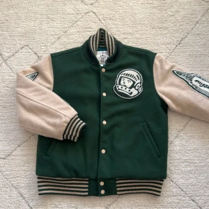 Grön och beige bomberjacka från Billionaire Boys Club - Snygg bomberjacka i grönt och beige med broderade detaljer från Billionaire Boys Club. Jackan har långa ärmar, ribbade muddar och en quiltad insida. På ryggen står 'HEART MIND' med ett hjärta. Perfekt för en cool och avslappnad stil.