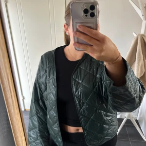 Grön quiltad jacka - Säljer en snygg grön quiltad jacka med dragkedja. Jackan har en modern design med långa ärmar och en lätt glansig yta. Perfekt för att hålla stilen under kyligare dagar. Passar bra till både casual och mer uppklädda tillfällen.
