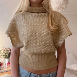 Beige stickad topp/poncho - Säljer en beige stickad topp med korta ärmar och hög krage. Toppen har en ribbad nederkant som ger en snygg passform. Perfekt för en stilren look.