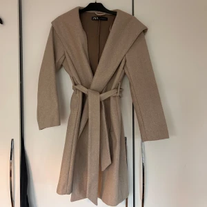 Beige kappa från Zara - Säljer en stilren beige kappa från Zara med huva och knytskärp i midjan. Kappan har långa ärmar och en elegant design som passar perfekt för kyligare dagar.