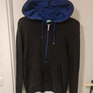 Svart hoodie från Boss - Säljer en stilren svart hoodie från Boss med blå insida i huvan och dragkedja. Perfekt för en avslappnad stil. Den har en klassisk känguruficka och justerbara snören i huvan.
