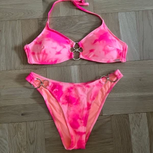 Rosa bikini med ringdetaljer - Trendig rosa bikini med tie-dye mönster och snygga ringdetaljer både på toppen och trosorna. Perfekt för sommarens stranddagar. Toppen har knytband i nacken för justerbar passform. Som en xs-s. Aldrig använd 
