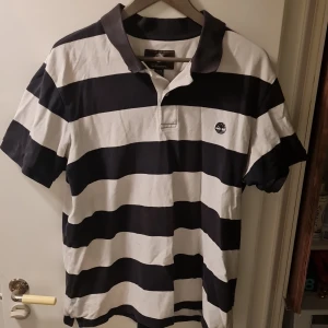 Randig pikétröja från Timberland - Säljer en klassisk svartvit randig pikétröja från Timberland. Tröjan har korta ärmar och en krage med knappar. Perfekt för en avslappnad stil.