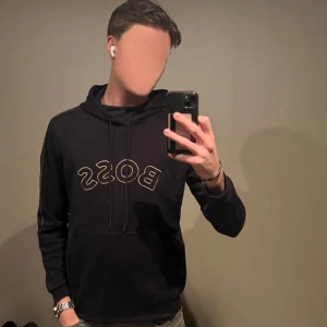 Svart hoodie från Boss - Säljer en stilren svart hoodie från Boss med deras logga i guld på bröstet. Tröjan har svarta detaljer på fickan som ger tröjan lite mer karaktär. Tröjan är i bra skick och det finns inga defekter på den. Hör bara av dig om du har frågor! 😄👍