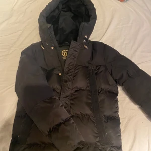 Svart pufferjacka från Glorious Gangsta - Säljer en svart pufferjacka från Glorious Gangsta med huva och dragkedja. Jackan har en stilren design med en logotyp på ärmen. Perfekt för kyliga dagar.