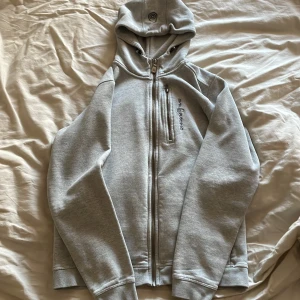 Grå huvtröja från Sail Racing - Säljer en stilren grå huvtröja från Sail Racing. Tröjan har dragkedja framtill och en praktisk huva med justerbar snörning. Perfekt för en avslappnad stil. 🧥