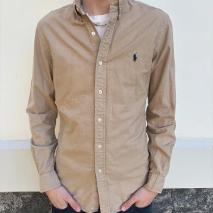 Polo Ralph Lauren skjorta - Säljer en stilren beige skjorta från Ralph Lauren i slim fit. Skjortan har långa ärmar och knappar framtill. Perfekt för en snygg och avslappnad look.