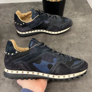 Valentino Rockrunner sneakers i blått och svart - Snygga Valentino Rockrunner sneakers i en cool blå och svart camouflage-design. Skorna har en unik kombination av mocka och syntetmaterial med ikoniska nitar runt sulan. Perfekta för att ge din stil en edgy touch.