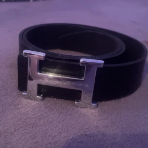 Svart bälte med silverfärgat spänne - Snyggt svart bälte med ett stort, silverfärgat H-format spänne. Perfekt för att ge en stilren touch till din outfit. Passar både till jeans och kostymbyxor.