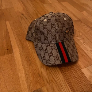 Beige keps med monogrammönster - Snygg beige keps  med klassiskt monogrammönster och röd-gröna ränder framtill. Perfekt för att ge din outfit en lyxig touch. Justerbar passform för optimal komfort.
