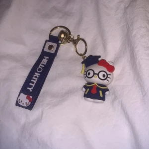 Hello Kitty nyckelring med studenttema - Charmig Hello Kitty nyckelring med en figur klädd i studentkläder och glasögon. Den har en blå rem med texten 'HELLO KITTY' och en gulddetalj. Perfekt som accessoar eller present!