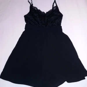 Svart spetsprydd jumpsuit - Snygg svart jumpsuit med spetsdetaljer upptill och smala axelband. Perfekt för en stilren look. Den har en elastisk midja för en bekväm passform.