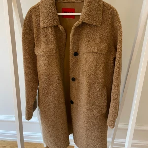 Beige teddyjacka från Hugo Boss - Säljer en snygg beige teddyjacka från Hugo Boss. Jackan har en klassisk krage och knäpps med svarta knappar framtill. Perfekt för kyligare dagar, vår och höst med sin mysiga och varma och tidlösa design.