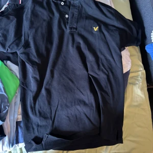 Svart pikétröja med logotyp - Säljer en klassisk svart pikétröja från Lyle & Scott med deras ikoniska logotyp på bröstet. Tröjan har en knappslå med två knappar och en bekväm passform. Perfekt för en stilren look.