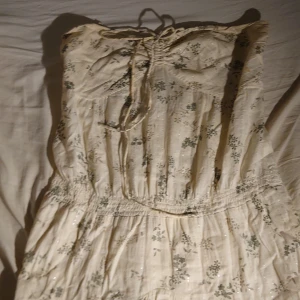 Blommig klänning från Vila - Charmig blommig klänning från Vila i en ljus färgton med gröna blommönster. Klänningen har en smockad överdel och spetsdetaljer vid fållen. Perfekt för sommarens varma dagar. Sorry att bilderna är sämst men jag har inga bilder med den på. Snören som man knyter runt axlarna saknas men det går att använda vilket snöre som helst, eller ha på sig den utan snören.