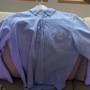 Blå skjorta från Gant - Säljer en stilren blå skjorta från Gant med en klassisk krage och knappar framtill. Skjortan har en bröstficka med Gant-logotypen broderad ovanför. Perfekt för en snygg och avslappnad look.