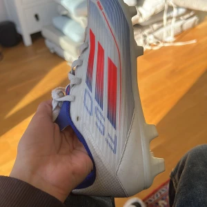 Fotbollsskor med röd och blå design - Snygga fotbollsskor med vit bas och röda samt blå detaljer. Skorna har snörning och är designade för optimalt grepp på planen med sina röda och vita dobbar.