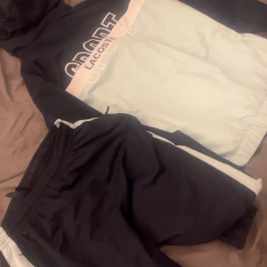 Svart och vit vindjacka från Lacoste - Säljer en stilren svart och vit vindjacka från Lacoste med dragkedja och hög krage. Jackan har långa ärmar och en sportig design med Lacoste-logga på bröstet. Perfekt för en aktiv livsstil.