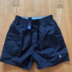 Svarta badshorts från Polo Ralph Lauren - Snygga svarta badshorts från Polo Ralph Lauren med elastisk midja och innerfoder i mesh. Perfekta för en stilren look vid stranden eller poolen. Klassisk logga broderad på benet.