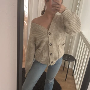 Beige stickad kofta - Säljer en mysig beige stickad kofta med stora knappar framtill. Koftan har en v-ringad design och långa ärmar. Perfekt för en avslappnad stil.