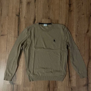 Beige tröja från Us Polo Assn - Säljer en stilren beige tröja från Us Polo Assn med rund hals och ett diskret broderat emblem på bröstet. Tröjan har långa ärmar och ribbade muddar vid ärmslut och nederkant. Perfekt för en klassisk och avslappnad look.