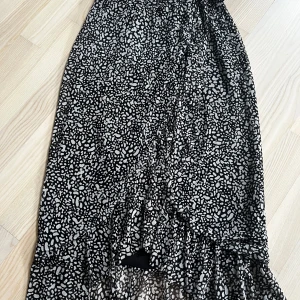 Svart och vit mönstrad kjol - Snygg lång omlottkjol med svart och vitt mönster. Kjolen har en volangdetalj längst ner som ger en fin rörelse. Perfekt för att ge en stilfull touch till din outfit.